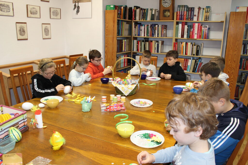 Grupa dzieci siedzi przy stole i przyozdabia styropianowe jajka.