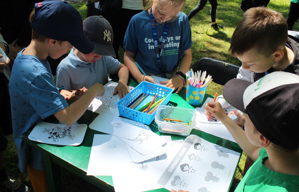 Na zdjęciu grupka dzieci rozwiązująca zagadki z motywami powieści Levisa Carrolla (malowanki, rebusy, labirynty).